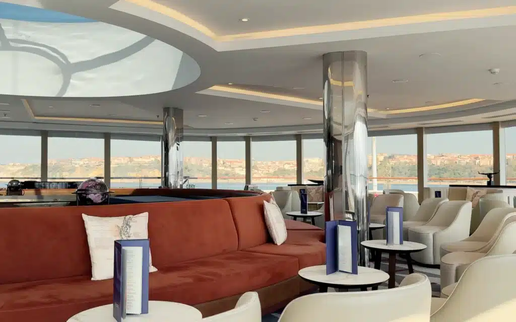 World Voyager Dome Observation Lounge interior showing rust-red curved velvet banquette seating, white armchairs, marble-topped side tables, blue cocktail menus, chrome columns, and wide panoramic windows framing coastal scenery