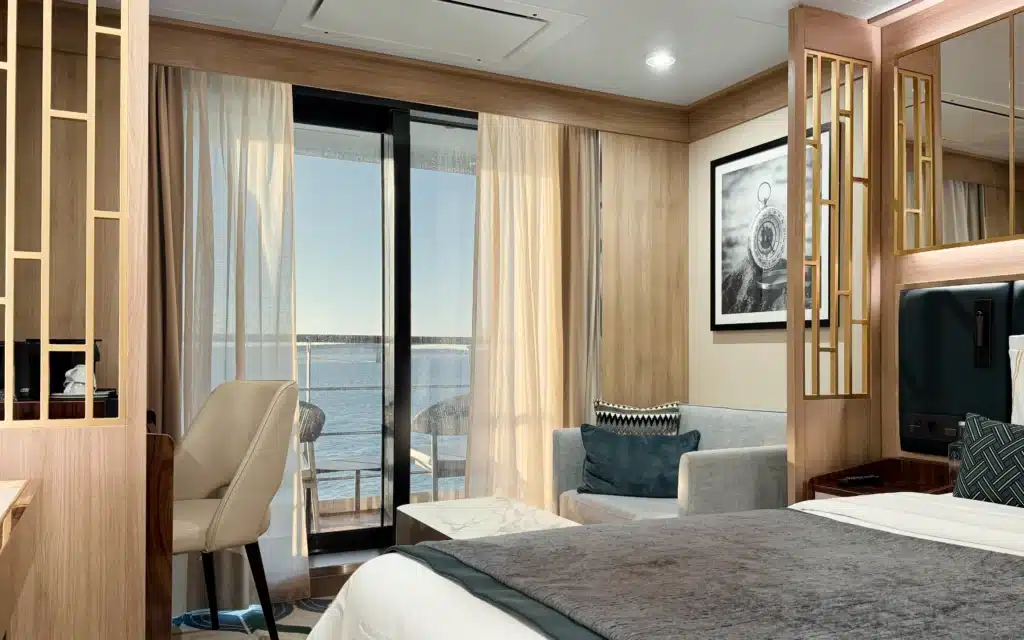 World Voyager Veranda Suite bedroom showing king bed, grey sofa, desk area, and sliding glass door opening onto a private veranda with ocean views