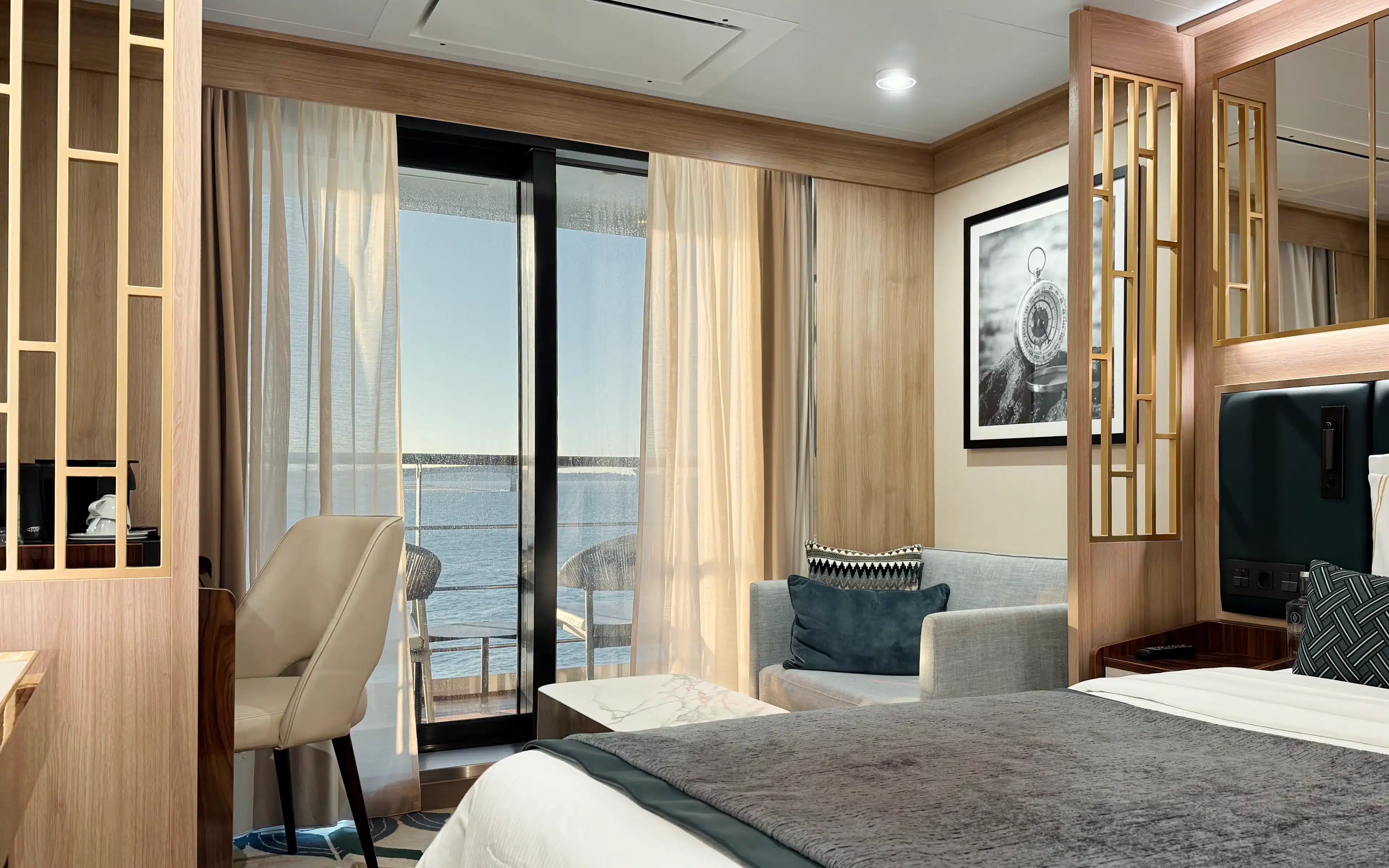 World Voyager Veranda Suite bedroom showing king bed, grey sofa, desk area, and sliding glass door opening onto a private veranda with ocean views