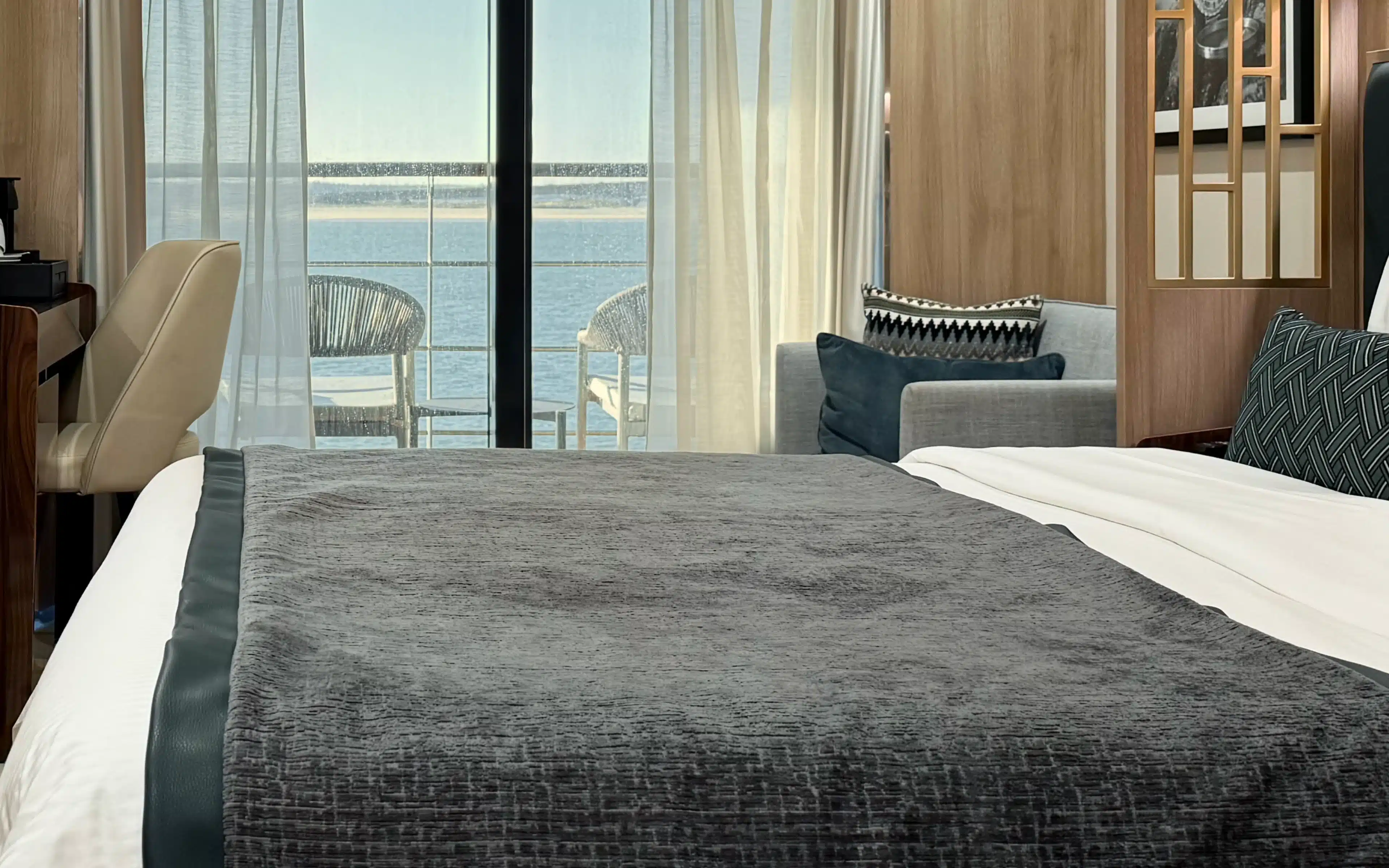 World Voyager Veranda Suite king bed in the foreground with grey throw, grey sofa sitting area, warm oak paneling, gold lattice screen detail, and a private veranda with rope chairs framing a sunlit coastal waterway beyond the sliding glass doors