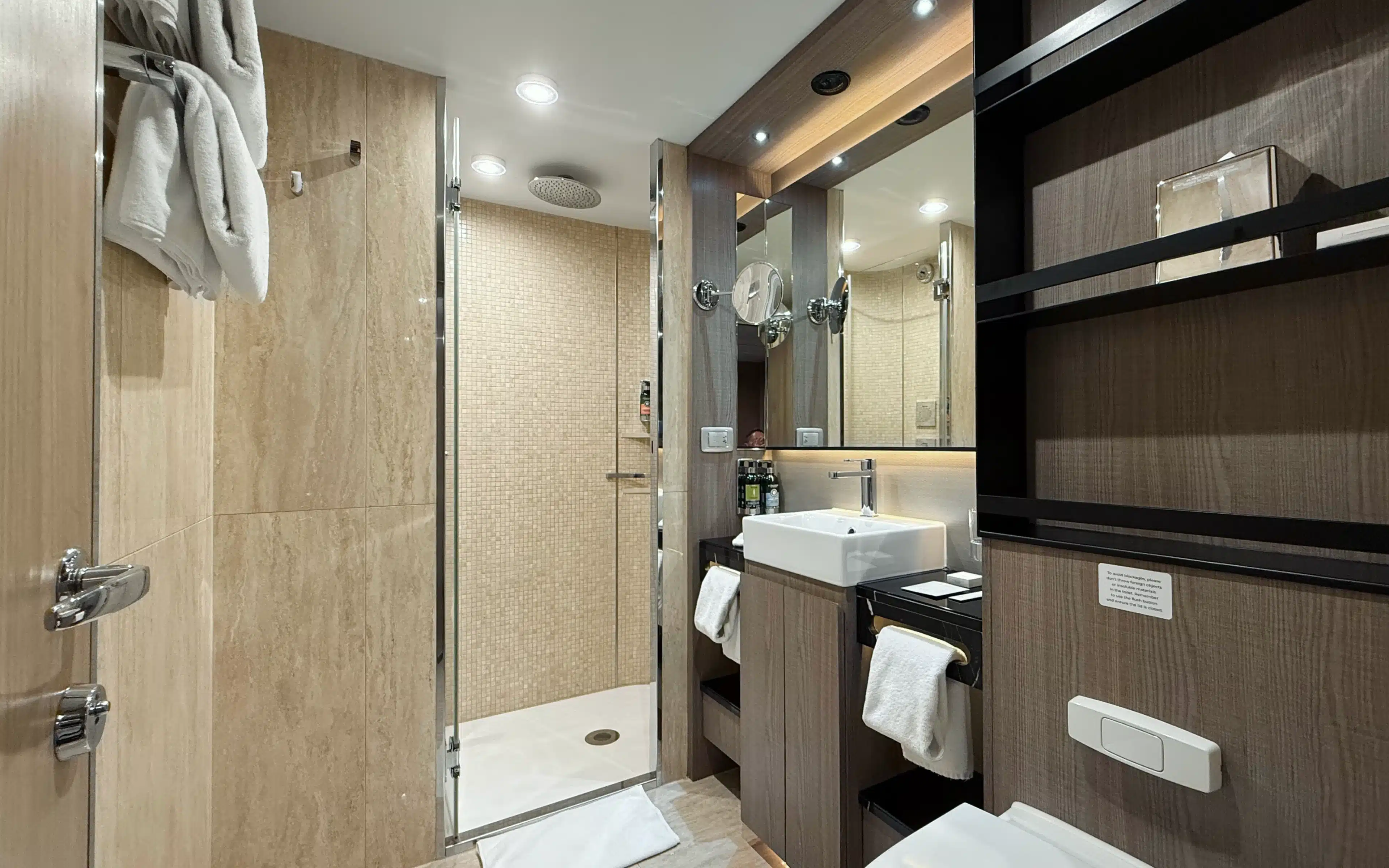 World Voyager Veranda Suite bathroom with spacious glass-enclosed shower, mosaic tile surround, rain head, vessel sink, and illuminated mirror with L'Occitane toiletries