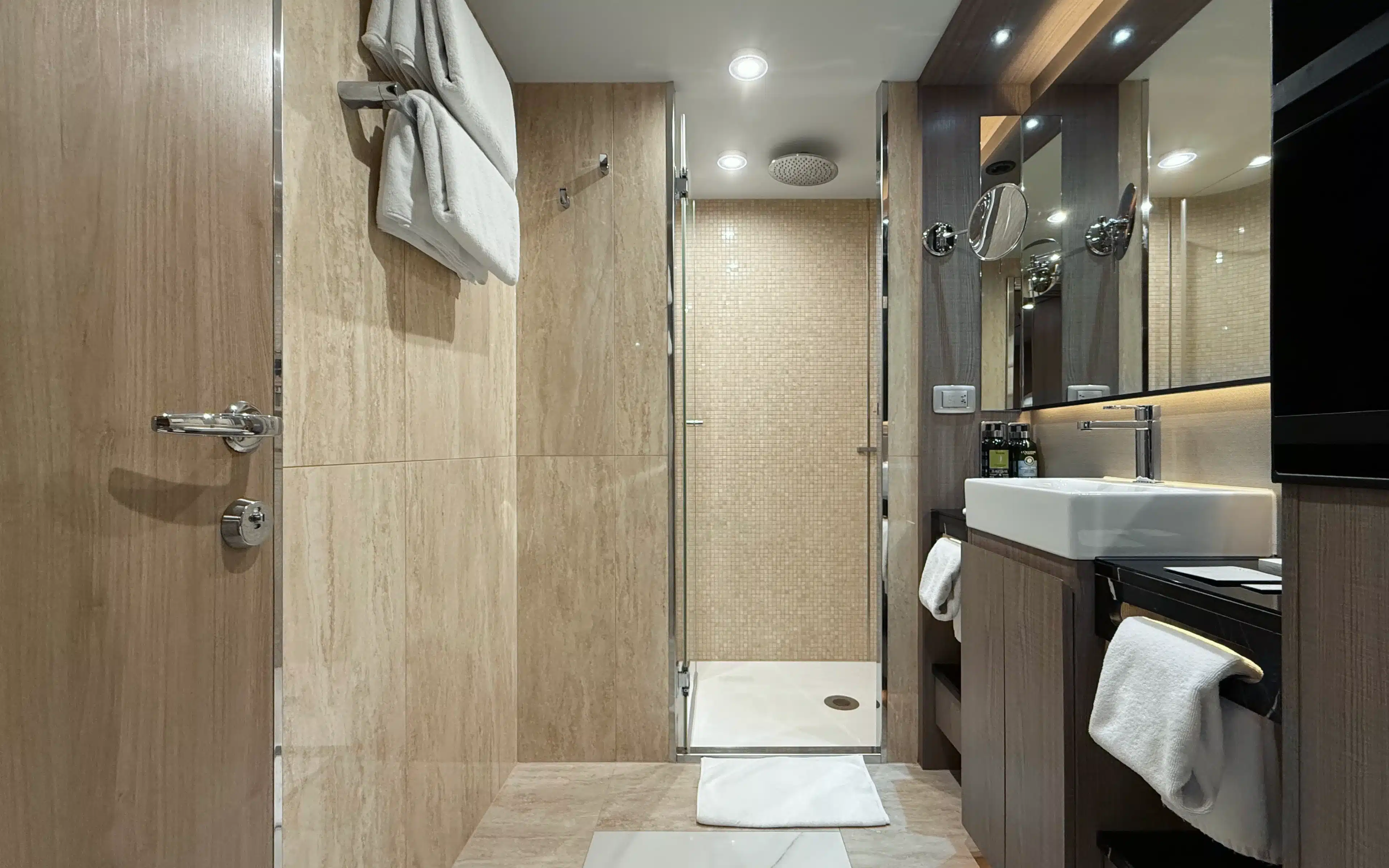 World Voyager suite bathroom featuring a generous glass-enclosed shower with full-height stone tile surround, rain head, and vessel sink with illuminated mirror