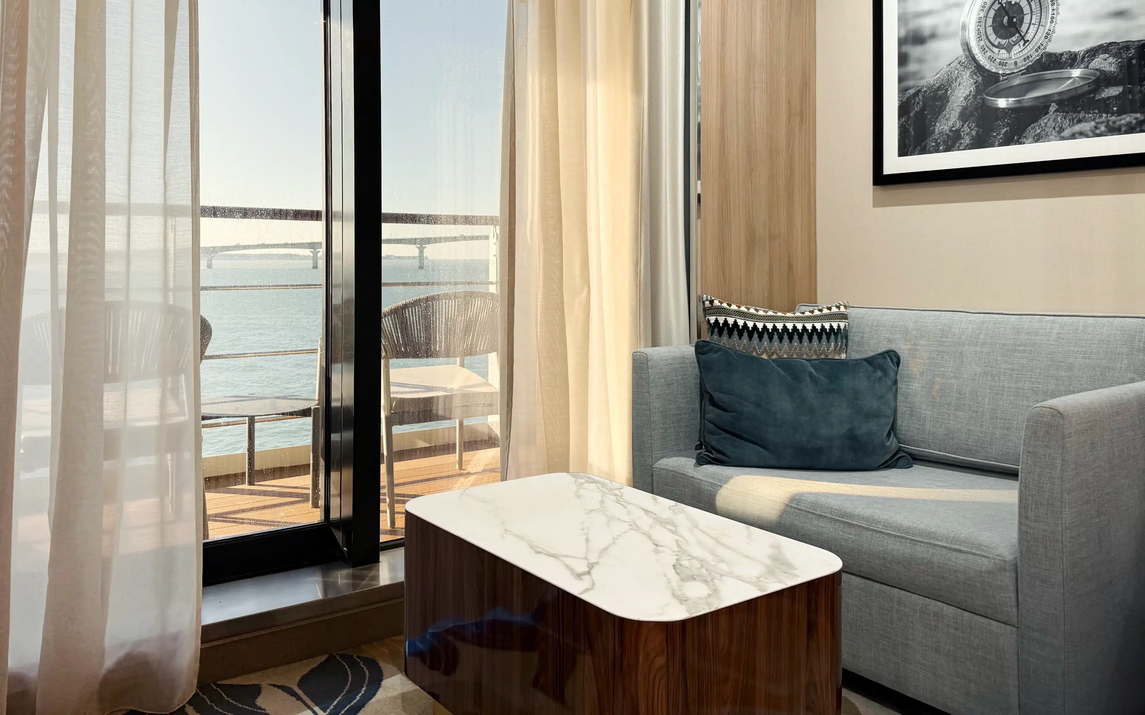 World Voyager Veranda Suite sitting area with grey two-seat sofa, marble-topped coffee table, and private veranda with ocean view beyond the sliding glass doors