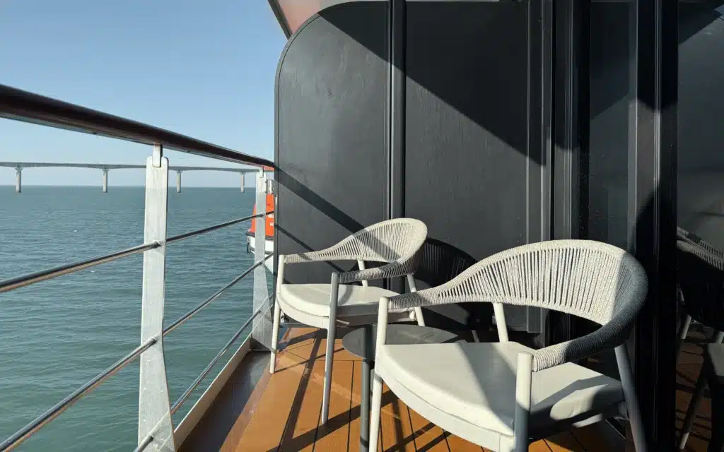 Private veranda of a World Voyager Veranda Suite with two woven rope chairs, teak decking, and a bridge visible over calm turquoise water