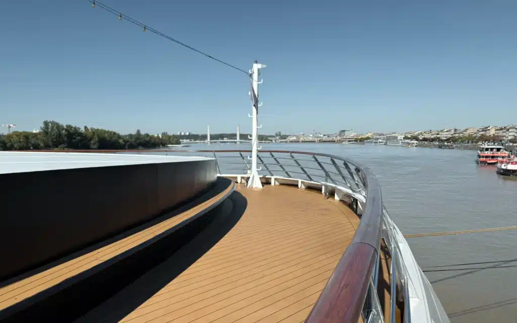 World Voyager Water's Edge forward observation deck showing curved teak decking sweeping around the bow, with the city of Bordeaux and the Garonne River visible beyond the polished railings under a clear blue sky