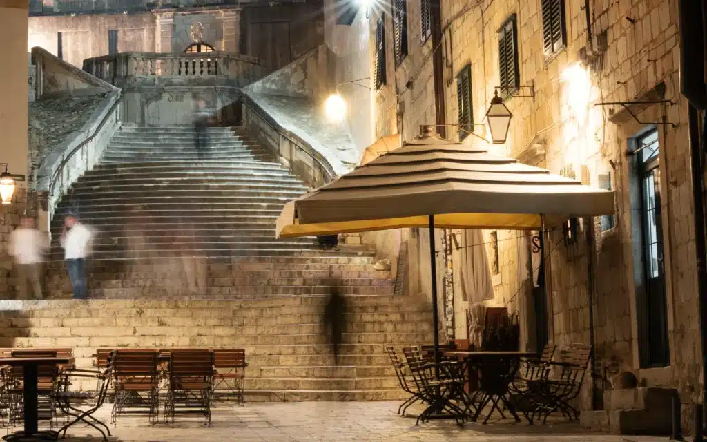 A long-exposure nighttime photograph of a cobblestone square in Dubrovnik's Old Town, with motion-blurred figures passing broad stone stairs rising to a Baroque church entrance, flanked by warm lantern-lit limestone buildings; a striped market umbrella shades bistro tables and wrought-iron chairs in the foreground