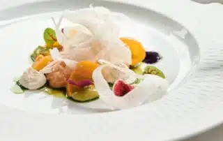 A fine-dining plate of dorado fillet presented on a wide white scallop-edged porcelain dish, garnished with shaved fennel, golden and green heirloom tomato segments, herb oil, small oyster mushrooms, micro sorrel leaves, and delicate pink and red edible flowers