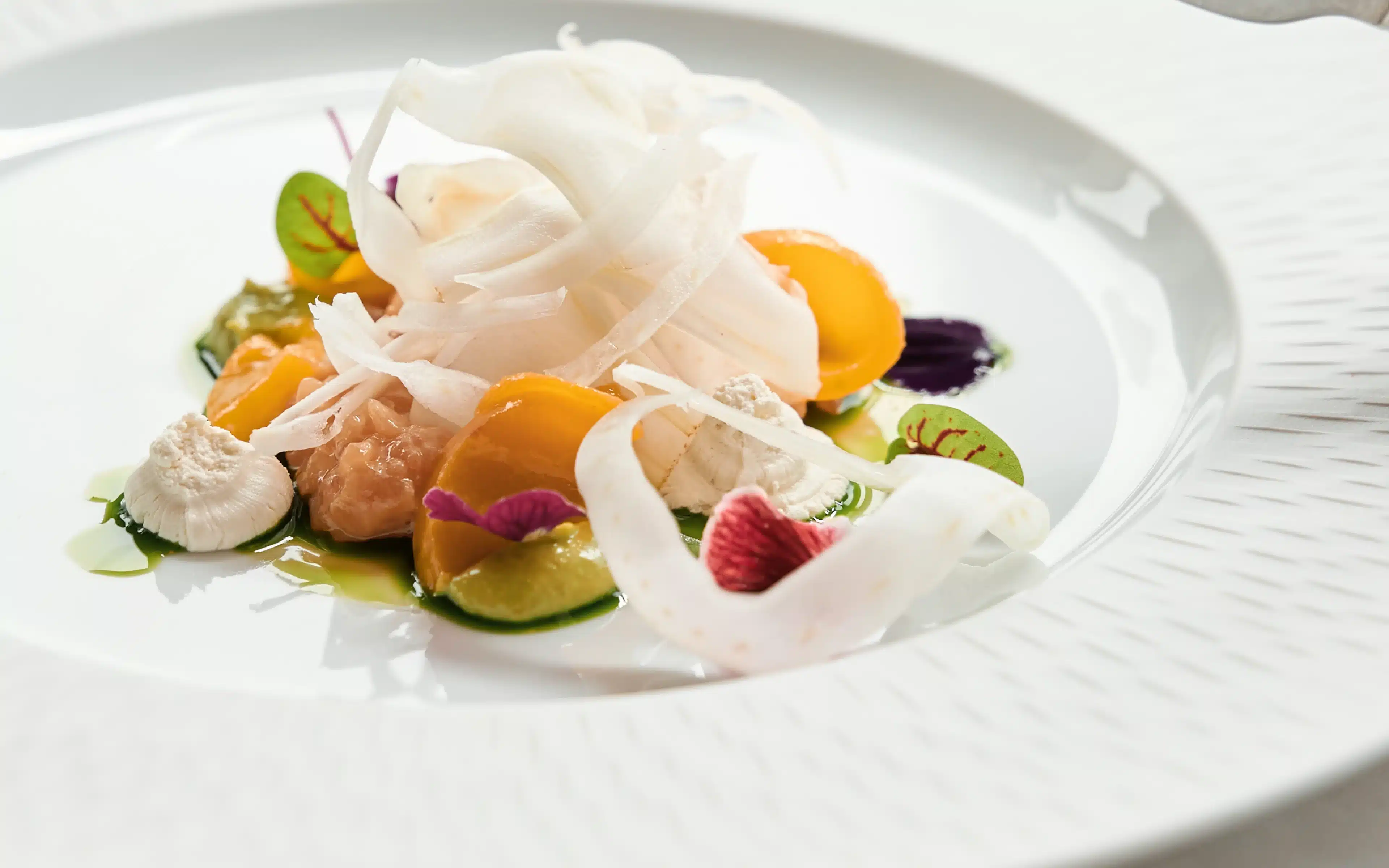 A fine-dining plate of dorado fillet presented on a wide white scallop-edged porcelain dish, garnished with shaved fennel, golden and green heirloom tomato segments, herb oil, small oyster mushrooms, micro sorrel leaves, and delicate pink and red edible flowers