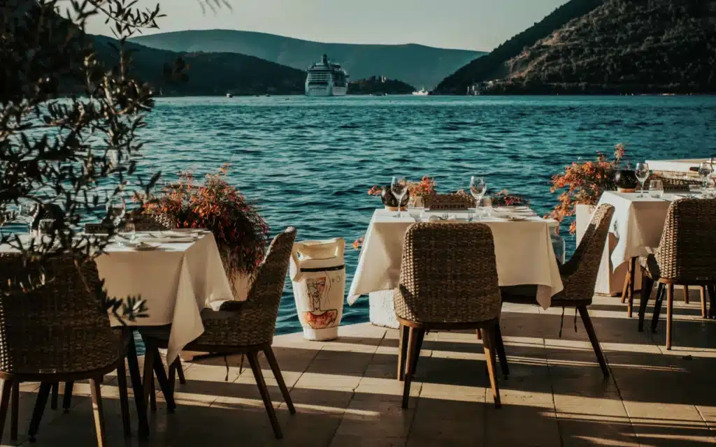 A waterfront restaurant terrace in Kotor, Montenegro, with white linen-draped tables set with wine glasses and orange floral arrangements in decorative ceramic urns, wicker and wood dining chairs, an olive tree in soft focus at left, and a large cruise ship anchored in the deep blue Bay of Kotor with forested mountains beyond