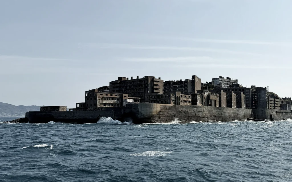 Abandoned concrete buildings and crumbling industrial structures crowd the fortified seawall of Hashima Island, known as Gunkanjima or Battleship Island, rising from choppy blue waters off Nagasaki, Japan, under a pale hazy sky
