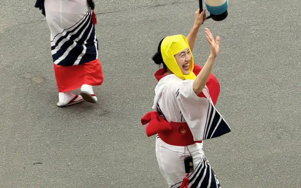 Aerial view of a joyful Japanese woman in traditional festival costume—white kimono with navy stripes, red obi sash, and yellow hood—dancing exuberantly in the street at Imabari, holding a small drum aloft with one hand raised, a second dancer in matching attire visible behind her