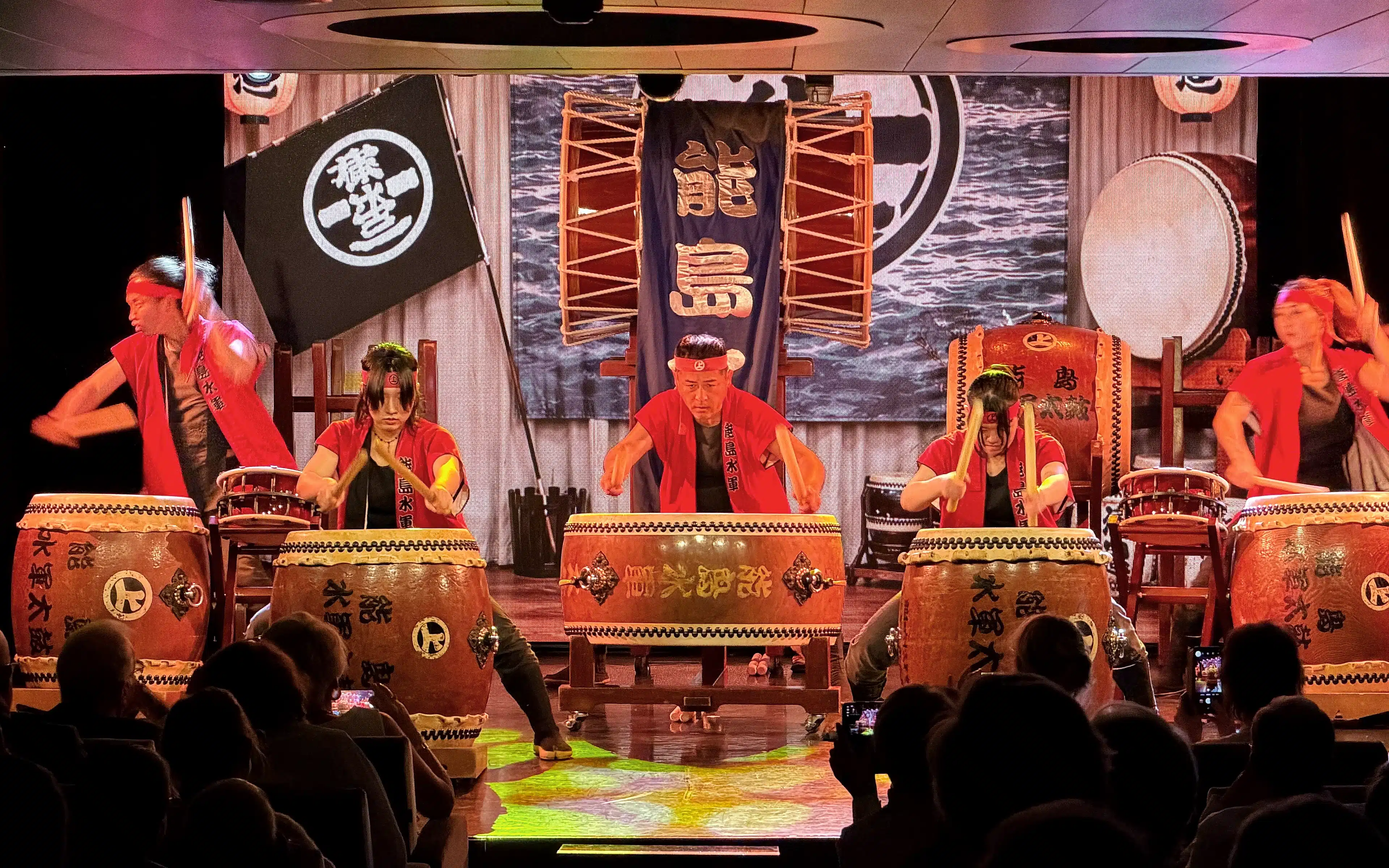 Five taiko drummers in red happi coats and black headbands perform with fierce energy on a cruise ship stage, striking large decorated barrel drums before an audience, with Japanese banners, lanterns, and a black clan flag displayed behind them