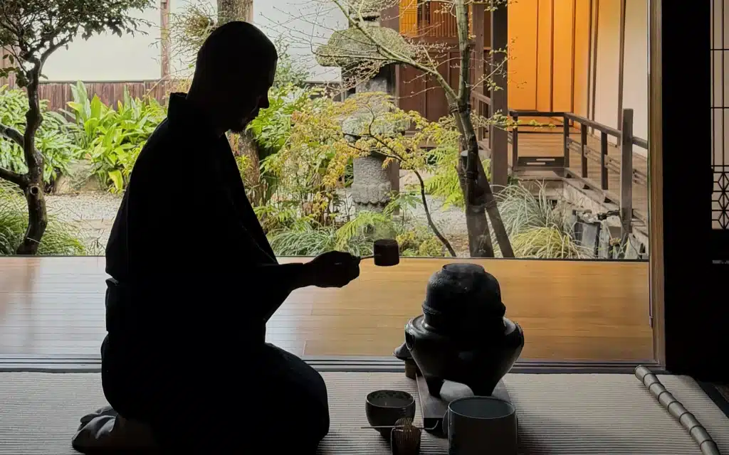 Silhouette of a man kneeling on tatami mats performing a traditional tea ceremony, holding a charcoal scoop above a black iron kettle and ceramic bowls, framed by an open doorway revealing a Japanese garden and timber building beyond