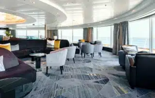 Observation Bar aboard Seabourn Quest after refurbishment, featuring a curved panoramic wall of floor-to-ceiling windows overlooking open ocean and the ship's deck railing, a sweeping dark upholstered banquette with gold and white accent cushions, gray fabric lounge chairs, a bold geometric patterned carpet in navy, gray, and gold, a sculptural layered oval ceiling detail, and floor-length gray drapes