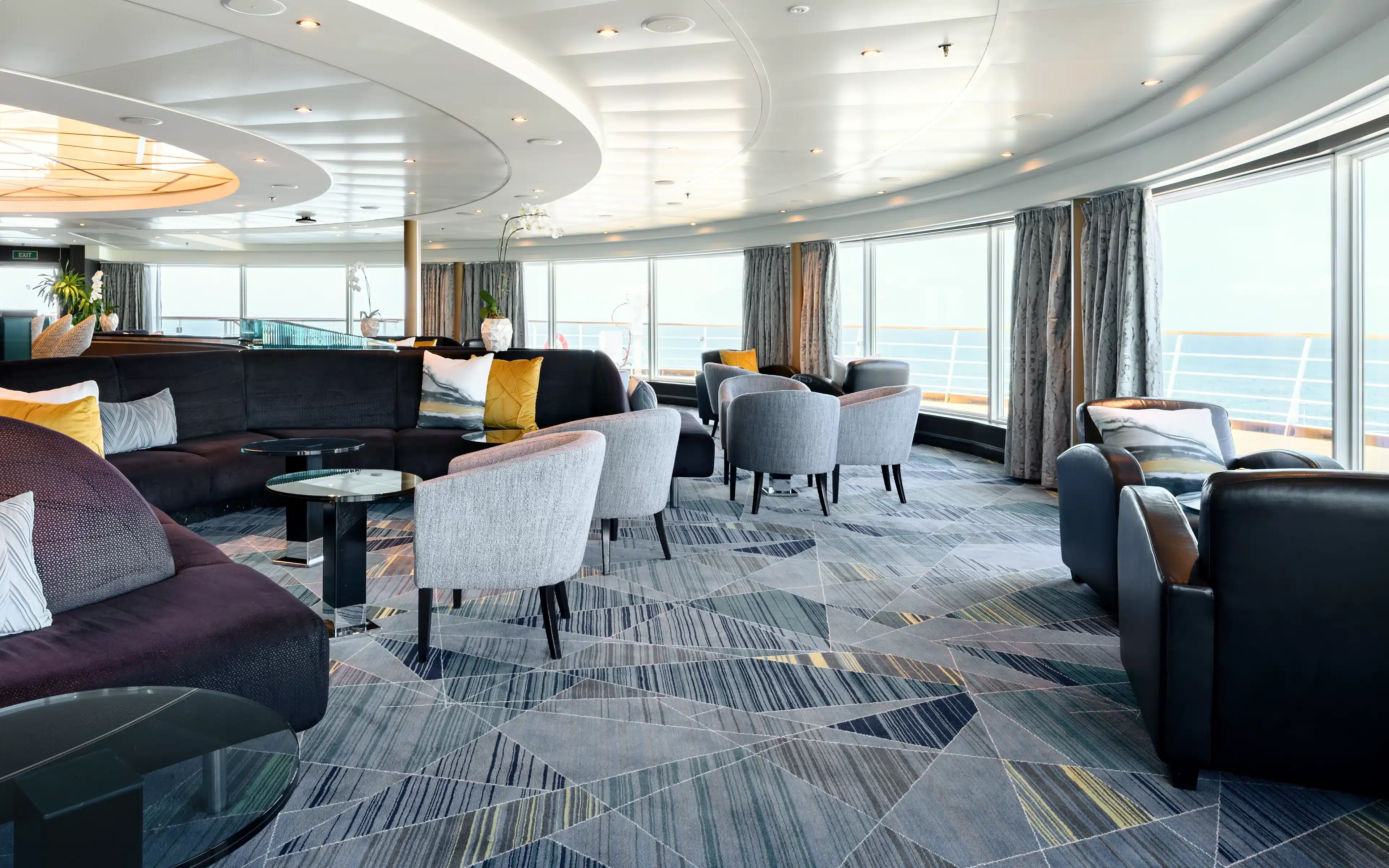 Observation Bar aboard Seabourn Quest after refurbishment, featuring a curved panoramic wall of floor-to-ceiling windows overlooking open ocean and the ship's deck railing, a sweeping dark upholstered banquette with gold and white accent cushions, gray fabric lounge chairs, a bold geometric patterned carpet in navy, gray, and gold, a sculptural layered oval ceiling detail, and floor-length gray drapes