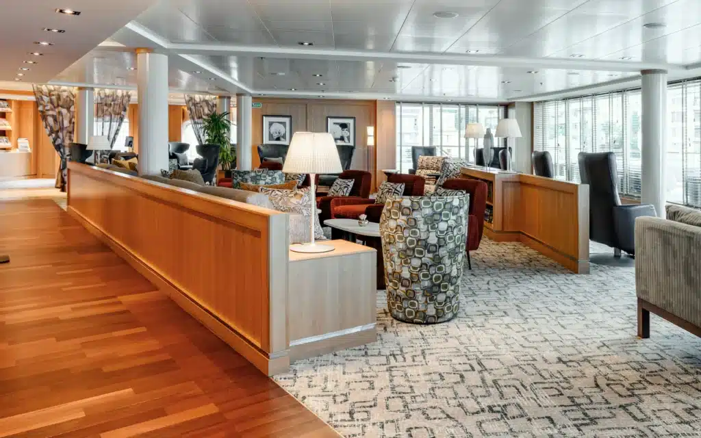 Seabourn Square aboard Seabourn Quest showing a wide, light-filled social lounge with warm honey-toned timber flooring and wall paneling, a central low divider, mixed seating including patterned barrel chairs, burgundy armchairs, and gray high-back chairs, white pleated table lamps, framed black-and-white portrait photography on the walls, potted greenery, and large windows with slatted blinds