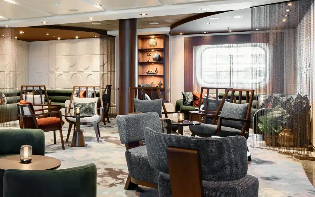 Refurbished lounge aboard Seabourn Quest featuring mid-century-style armchairs in charcoal boucle and burnt-orange velvet, deep green banquette seating, walnut-framed furniture, a lit display cabinet with decorative objects, patterned carpet in soft blue and gray tones, sheer metallic chain curtains, and recessed ceiling lighting