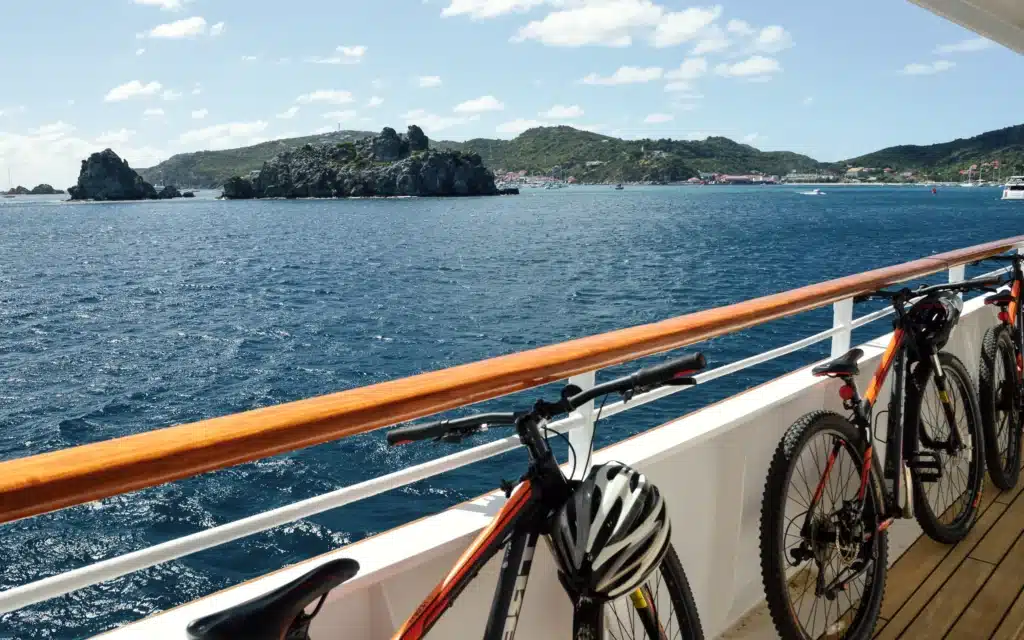 Three black and orange mountain bikes with helmets resting on handlebars lean against a white railing with a teak handrail on a SeaDream vessel deck, with rocky islets and the green hills of St. Barts visible across deep blue water