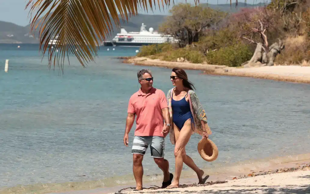 A couple walks hand-in-hand along a sandy Caribbean shoreline beneath a palm frond, with a SeaDream Yacht Club vessel anchored in the calm turquoise bay behind them
