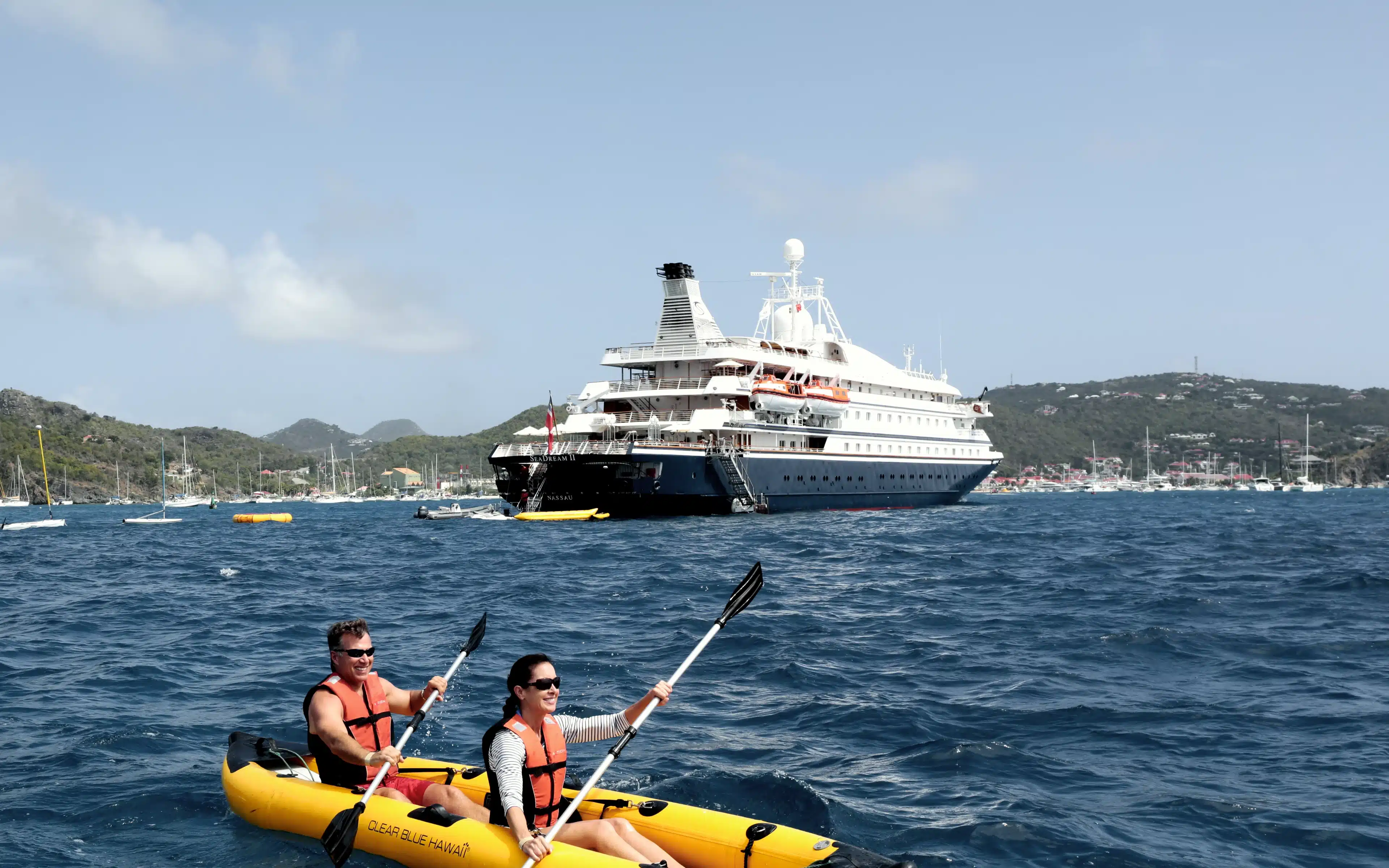 A man and woman in orange life jackets paddle a yellow inflatable tandem kayak on choppy open water, with SeaDream II anchored in the harbor at St. Barts and green hills beyond