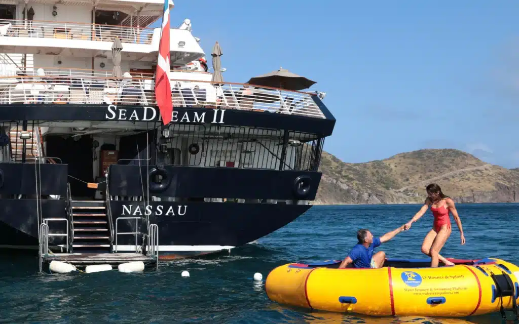 A woman in a red swimsuit steps onto a yellow inflatable water bouncer platform alongside the dark navy hull of SeaDream II, registered in Nassau, while a man assists her from the water