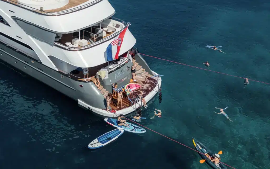 Aerial view of guests swimming, paddleboarding, and kayaking from the stern swim platform of Unforgettable Croatia's ship Aretis, anchored in vivid blue Adriatic waters