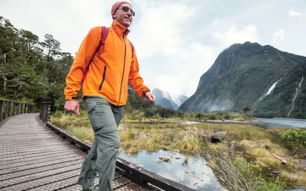 Hiker in an orange jacket crossing a boardwalk at Milford Sound with waterfalls and mountain peaks behind