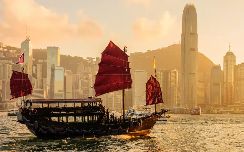 A red-sailed junk boat crosses Hong Kong's Victoria Harbour at sunset