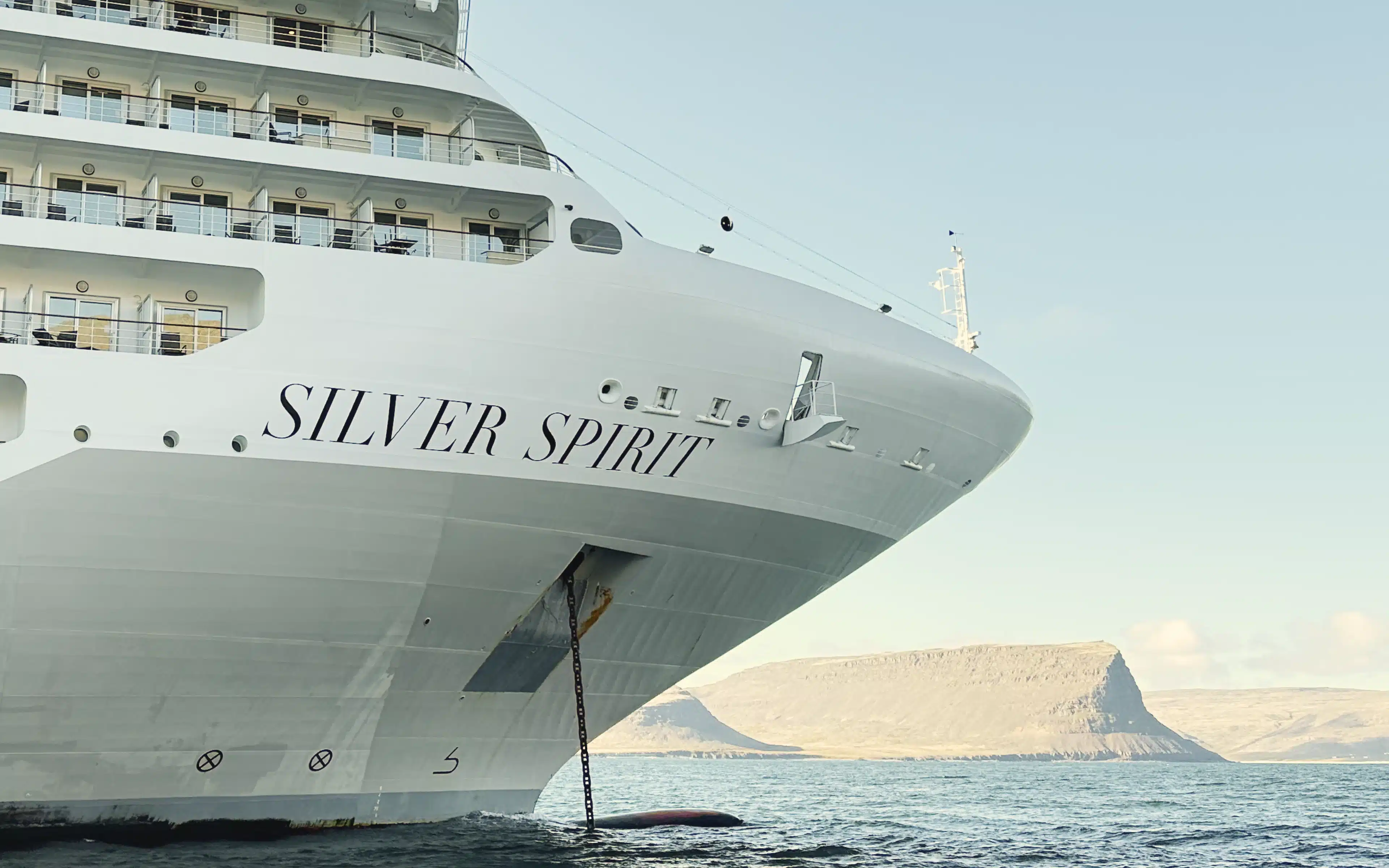 Bow of Silversea's Silver Spirit at anchor, its name in black script on the white hull, with a flat-topped headland rising across calm water beyond
