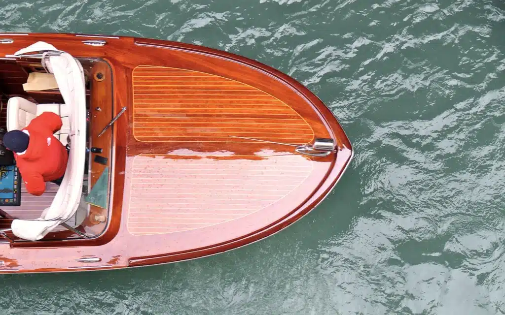 Overhead view of a varnished wooden motorboat with a cream-cushioned cockpit and a driver in a red jacket, moving across green water
