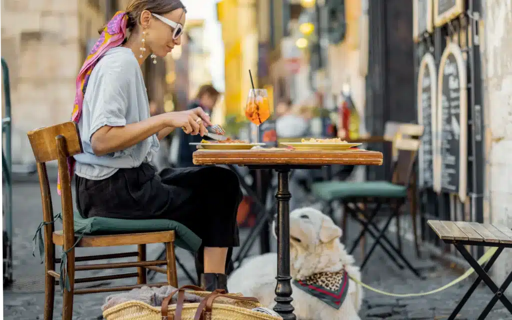 Woman in sunglasses and a patterned headscarf eating lunch at a small outdoor café table, a white dog in a bandana lying beneath it on a cobbled lane