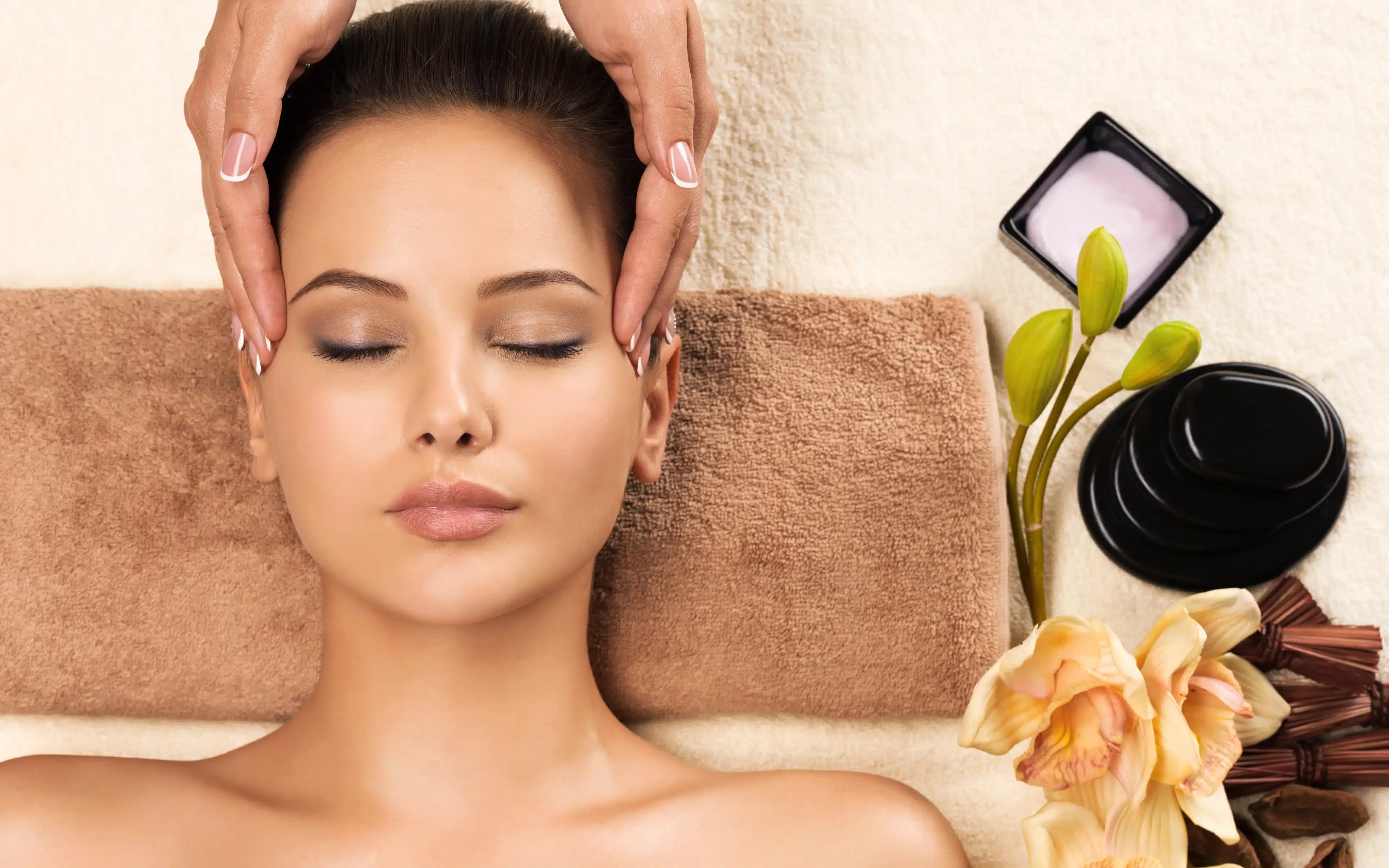Overhead view of a woman lying on a towel with her eyes closed as a therapist’s hands massage her temples, beside black massage stones, a dish of pink cream, orchids and green flower buds