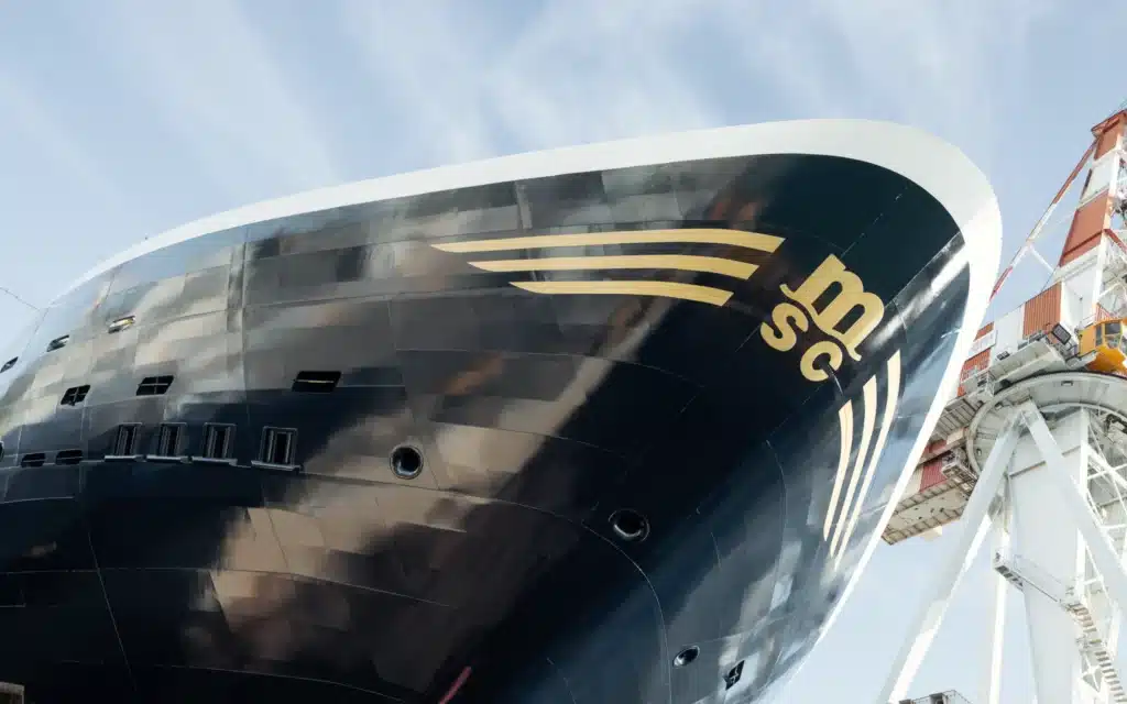 Black bow of an Explora Journeys ship bearing the gold MSC emblem, with a white shipyard crane and Fincantieri building against a pale blue sky