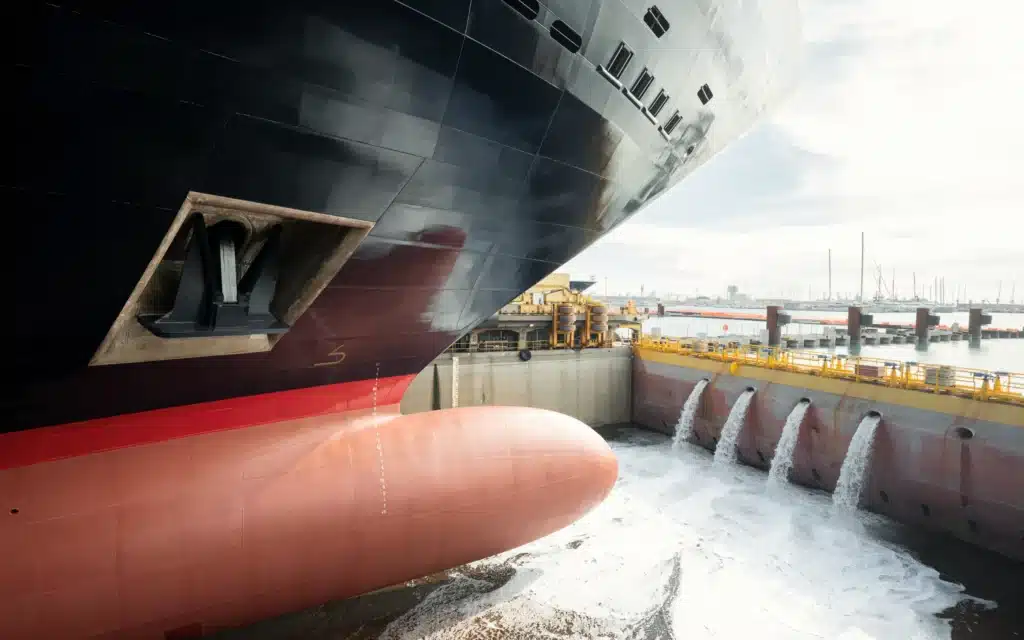 Low view of a ship’s bow and red bulbous bow in dry dock at Fincantieri Genoa as water pours in through four outlets, with a marina of yacht masts in the distance