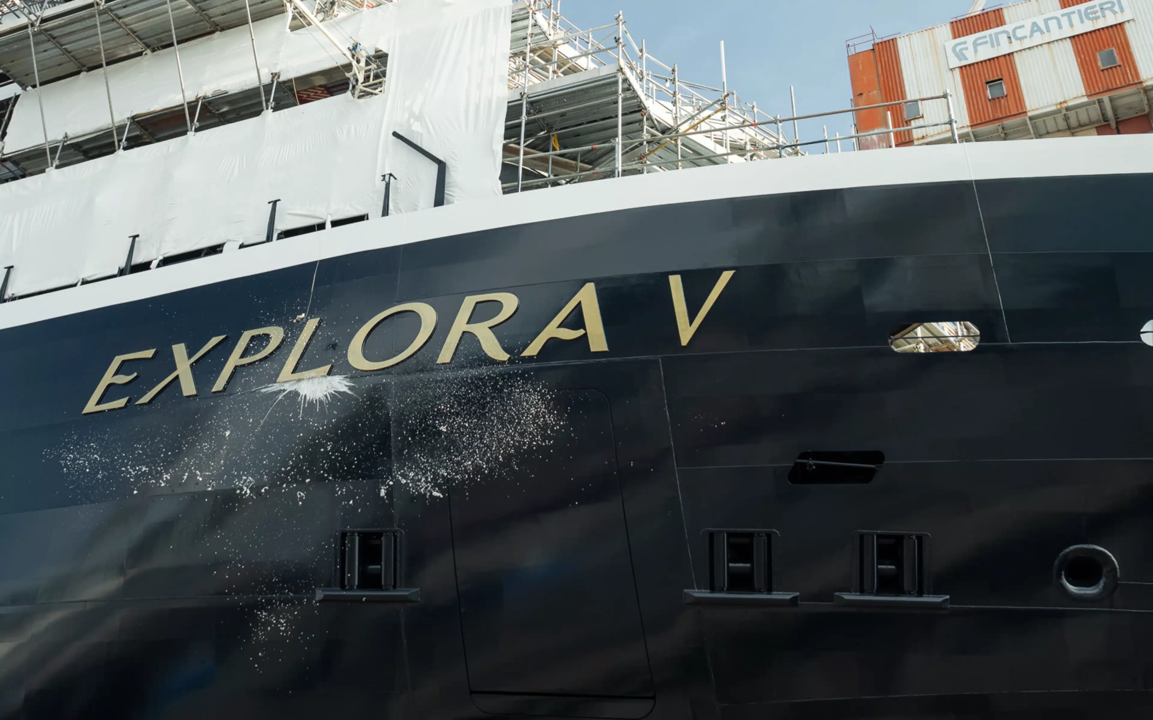 Gold “Explora V” lettering on the dark navy hull of Explora V, with splash marks on the paintwork, scaffolding and white sheeting on the upper decks and a Fincantieri building behind