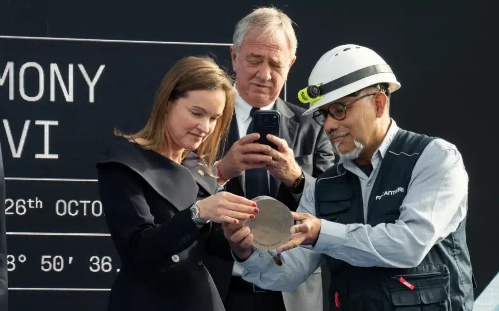 Anna Nash, President of Explora Journeys, holds the engraved Explora VI coin ceremony plaque with a Fincantieri worker in a hard hat, as Pierfrancesco Vago, Executive Chairman of MSC Group’s Cruise Division, photographs it on his phone