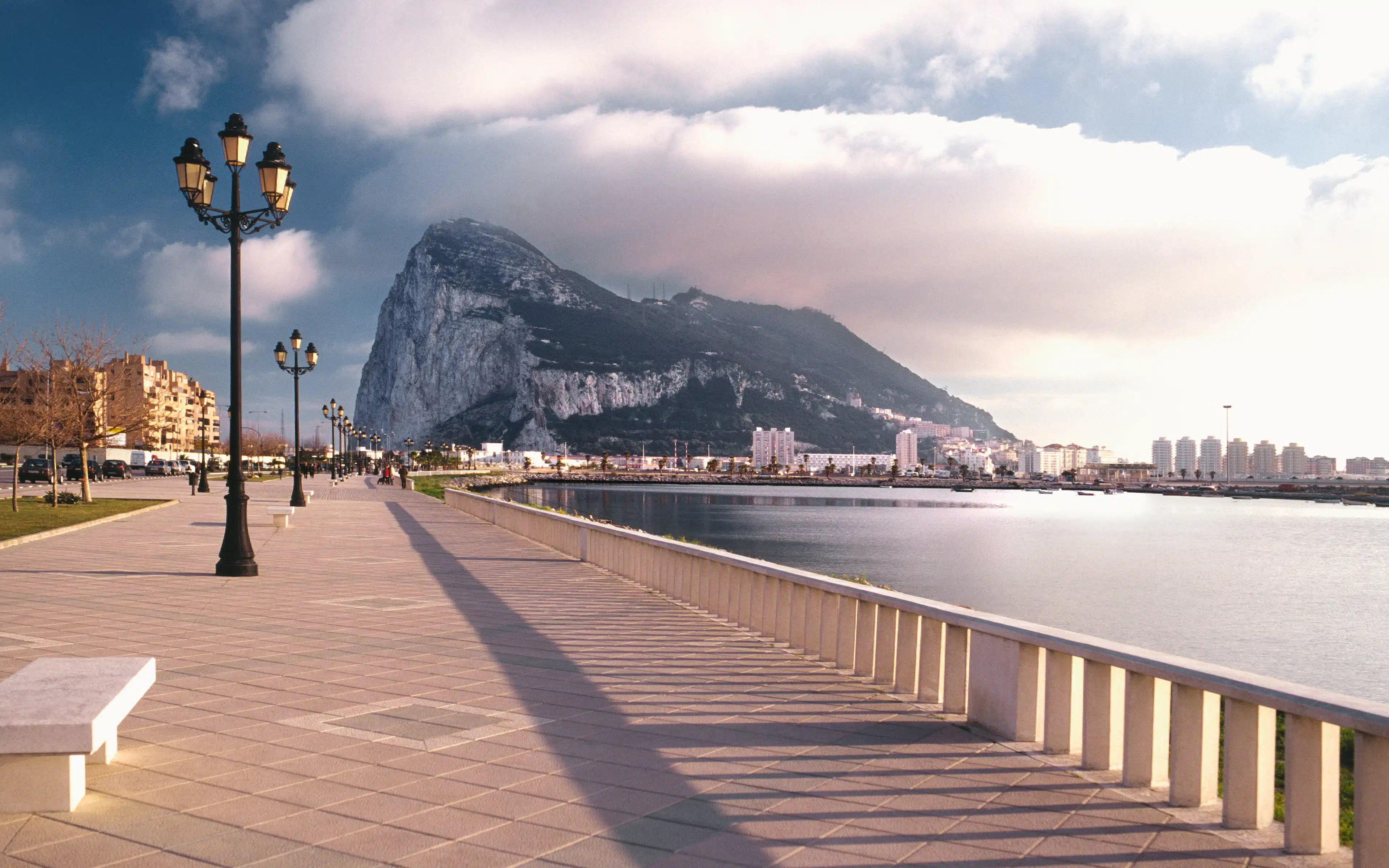 Seafront promenade with ornate lampposts looking toward the Rock of Gibraltar beneath dramatic clouds