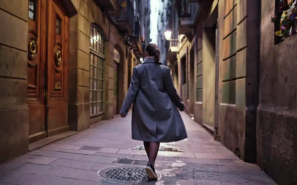 Woman in a long gray tweed coat walking along a narrow stone lane lined with old buildings and lamplight