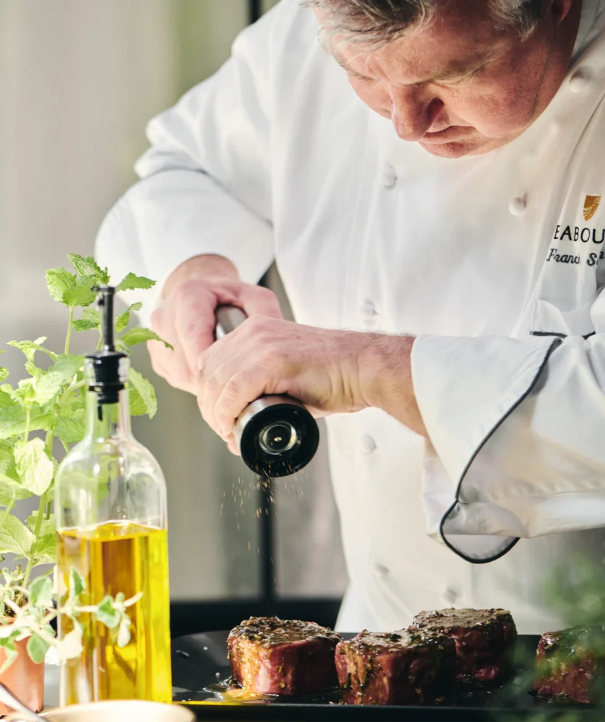 Seabourn chef in a white jacket grinding pepper over herb-marinated steaks beside a bottle of olive oil and fresh mint