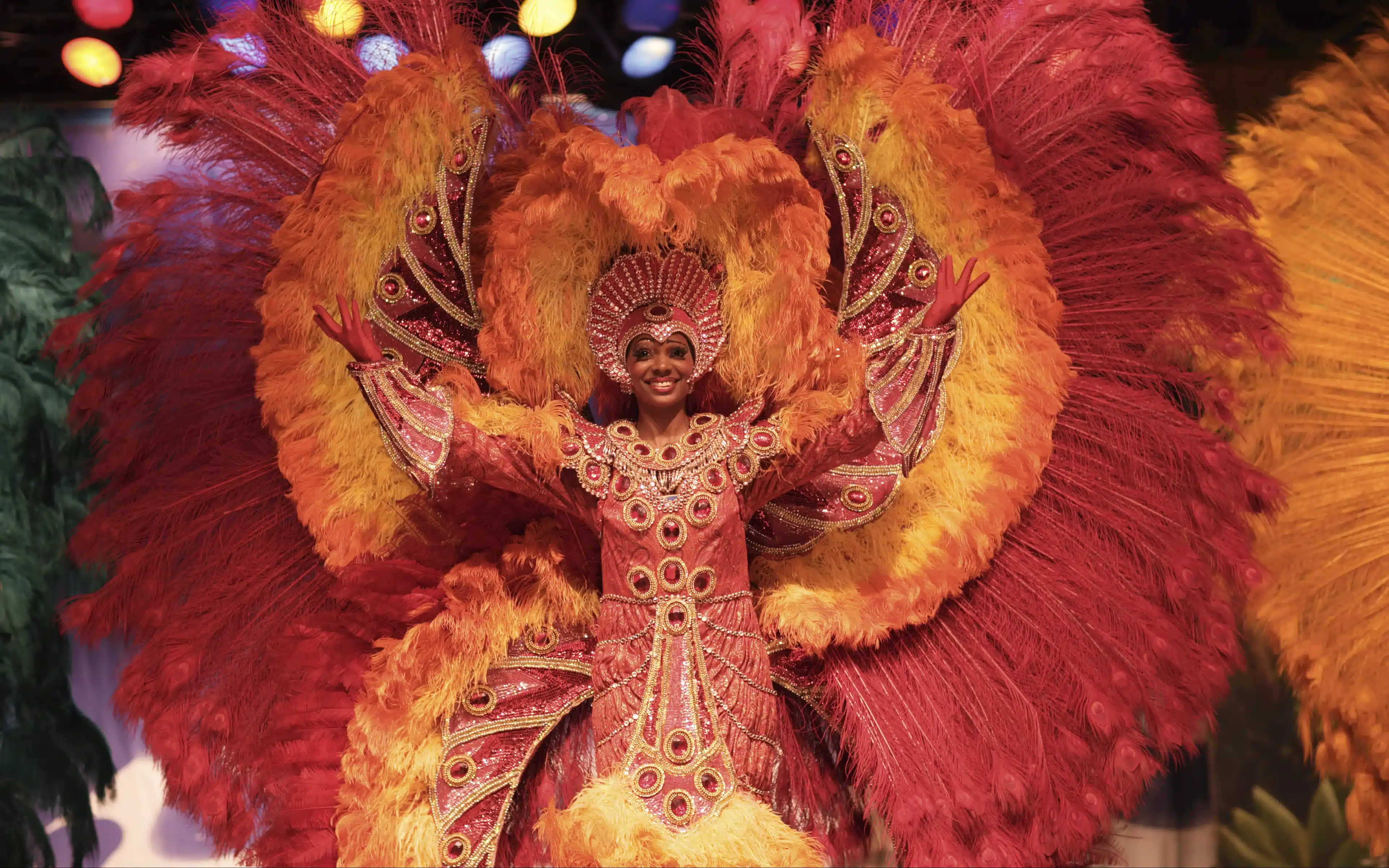 Smiling Carnival performer with arms raised in a jeweled red costume framed by huge red and orange feather plumes