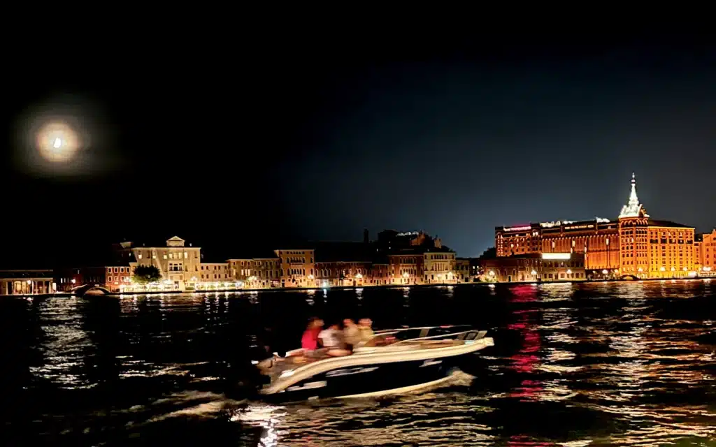Moonlit view across the Giudecca Canal in Venice at night, with the illuminated Molino Stucky building and a speedboat crossing the water