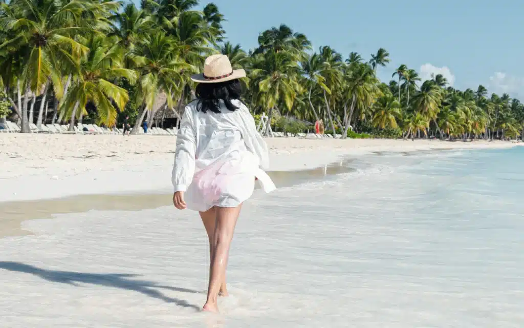 Woman in a straw hat and white shirt walking barefoot through shallow surf along a palm-lined white sand beach