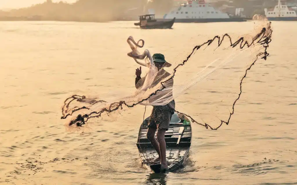 A fisherman standing in a small wooden boat casts a wide net across golden river water at sunset, with ferries in the background