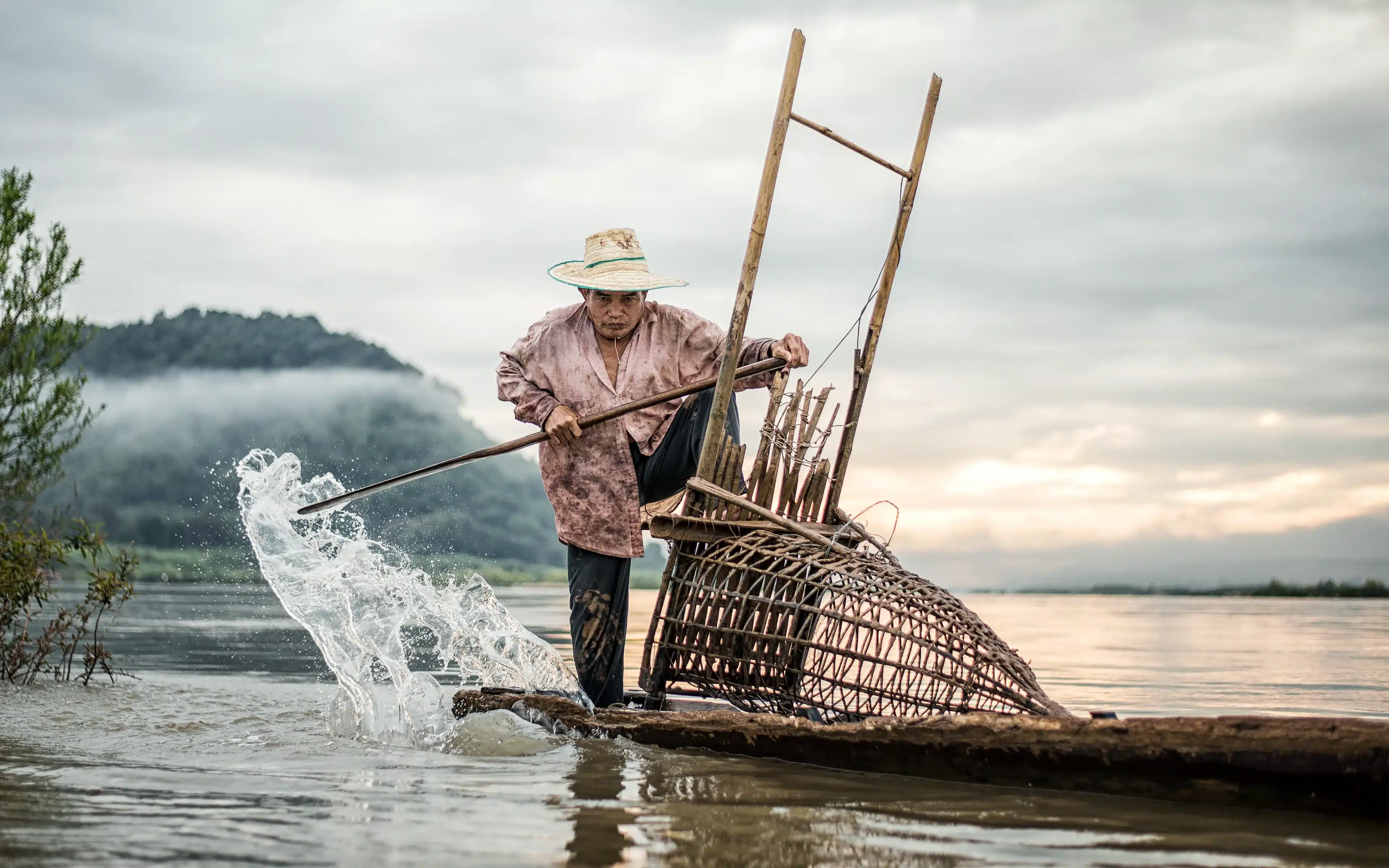 A fisherman in a straw hat stands on a wooden boat beside a large woven bamboo fish trap, splashing water with a paddle as mist drifts over a forested hill