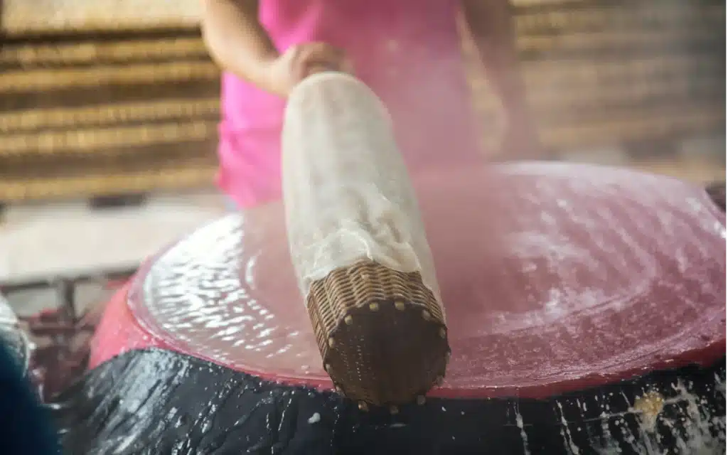 A woman in pink lifting a fresh sheet of rice paper from a steaming cloth on a woven bamboo roller