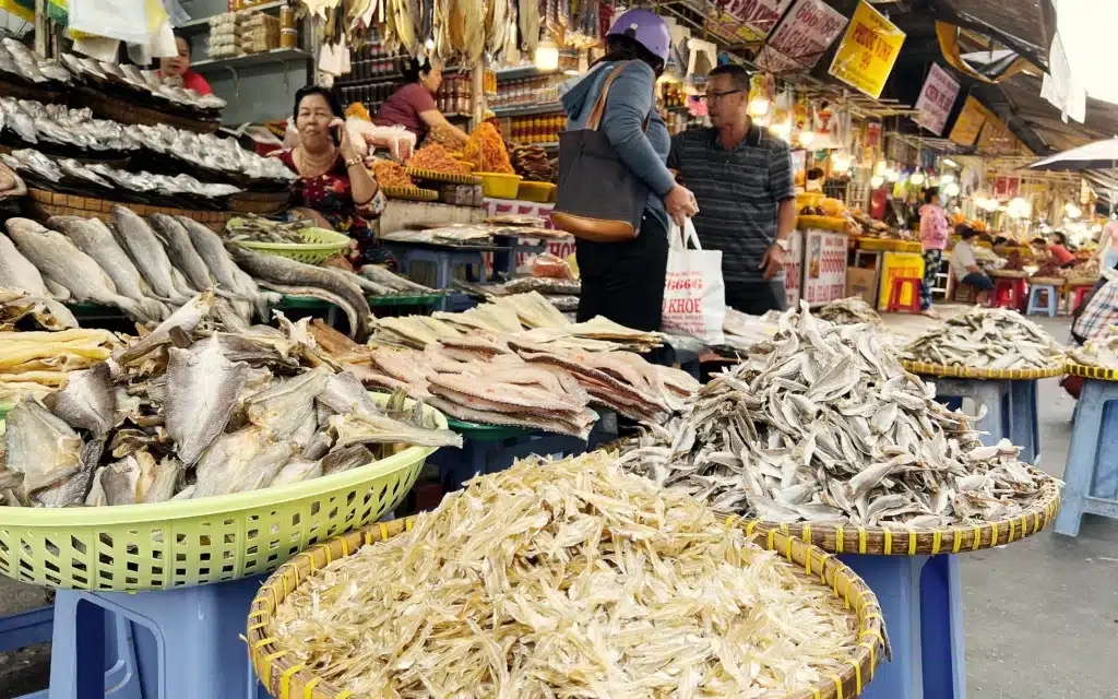 Baskets of dried fish and salted fillets stacked high at a busy Vietnamese market stall, with shoppers and vendors among yellow shop signs