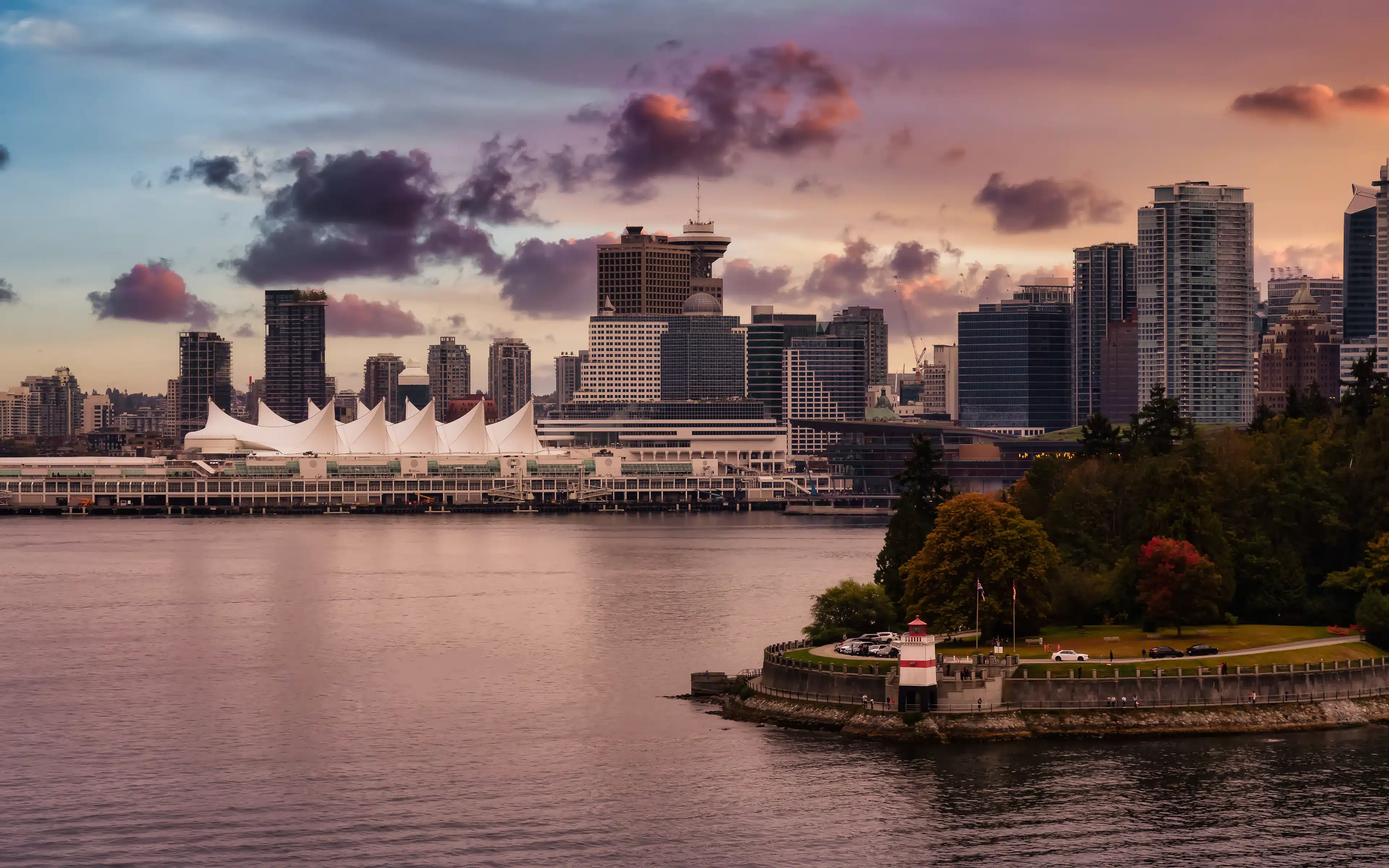 Canada Place cruise terminal and the downtown Vancouver skyline at dusk, with a red-and-white lighthouse on a wooded shoreline in the foreground
